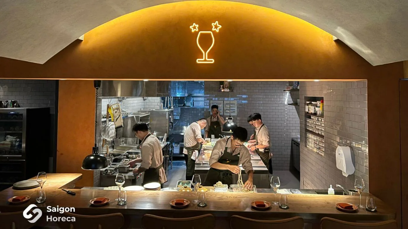 Không gian quầy bar gỗ sồi và ô vòm bếp mở nghệ thuật dưới đèn neon ly rượu vang tại Little Bear Thảo Điền
