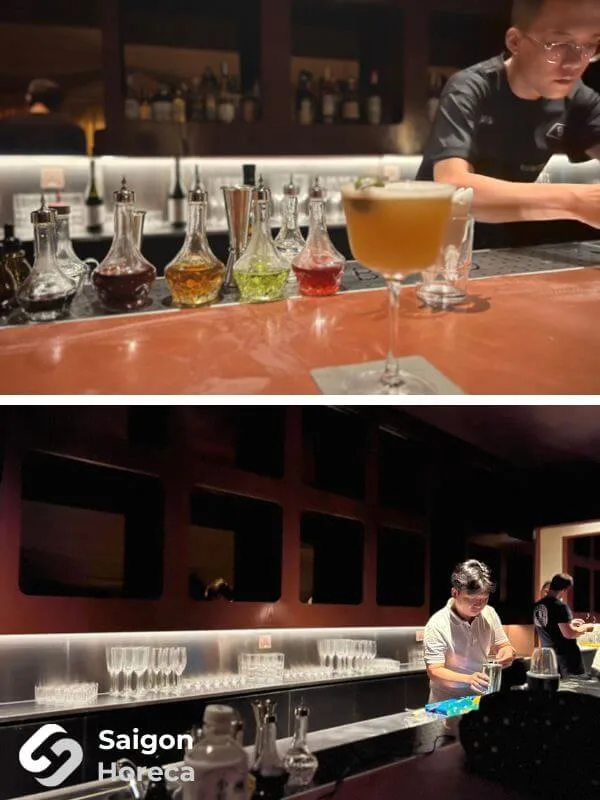 Bartender pha chế tại quầy bar bọc đồng với decanter và kệ ly back-bar tại Bambino