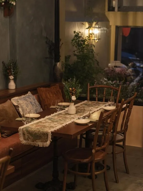 Góc ngồi ấm cúng với ghế băng gỗ, bàn trải khăn runner hoa – nơi khách thưởng thức bánh ngọt kiểu Âu