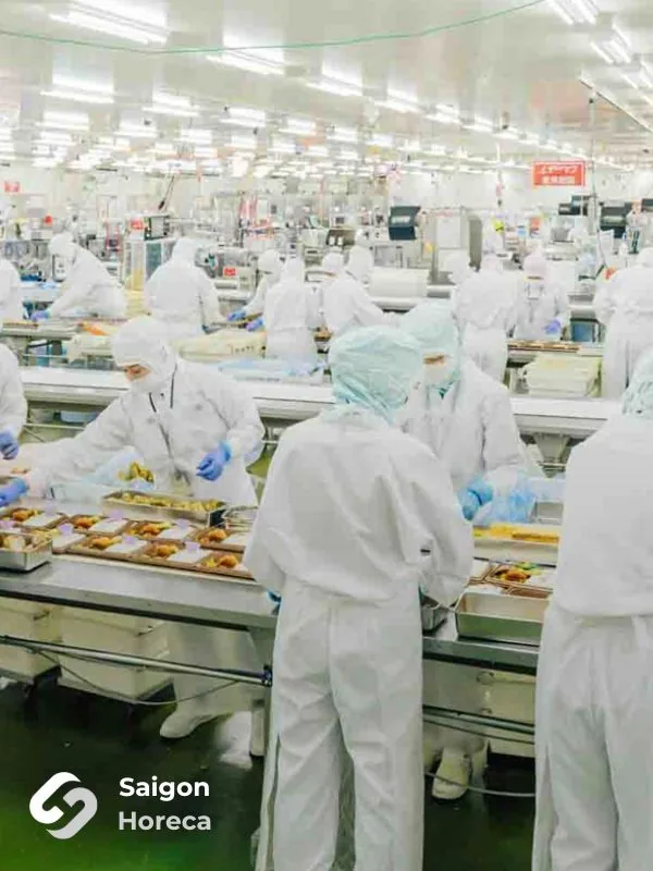 Công nhân mặc đồ bảo hộ xếp món chiên vào khay trên dây chuyền băng tải inox trong nhà xưởng chế biến thực phẩm Nichiyo