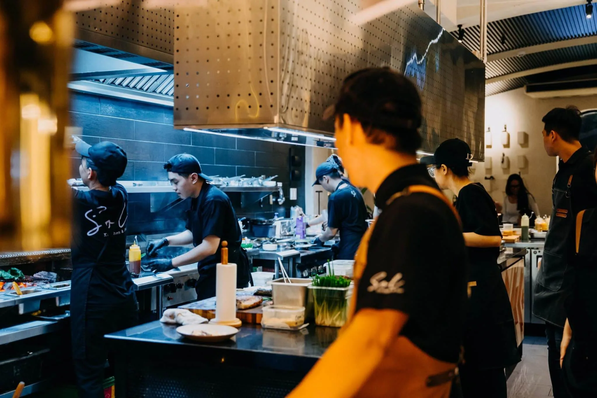 Khu bếp mở của nhà hàng Sol Kitchen & Bar với đầu bếp mặc đồng phục đen tạp dề cam đang nấu tại dãy bếp inox và chụp hút khói thép